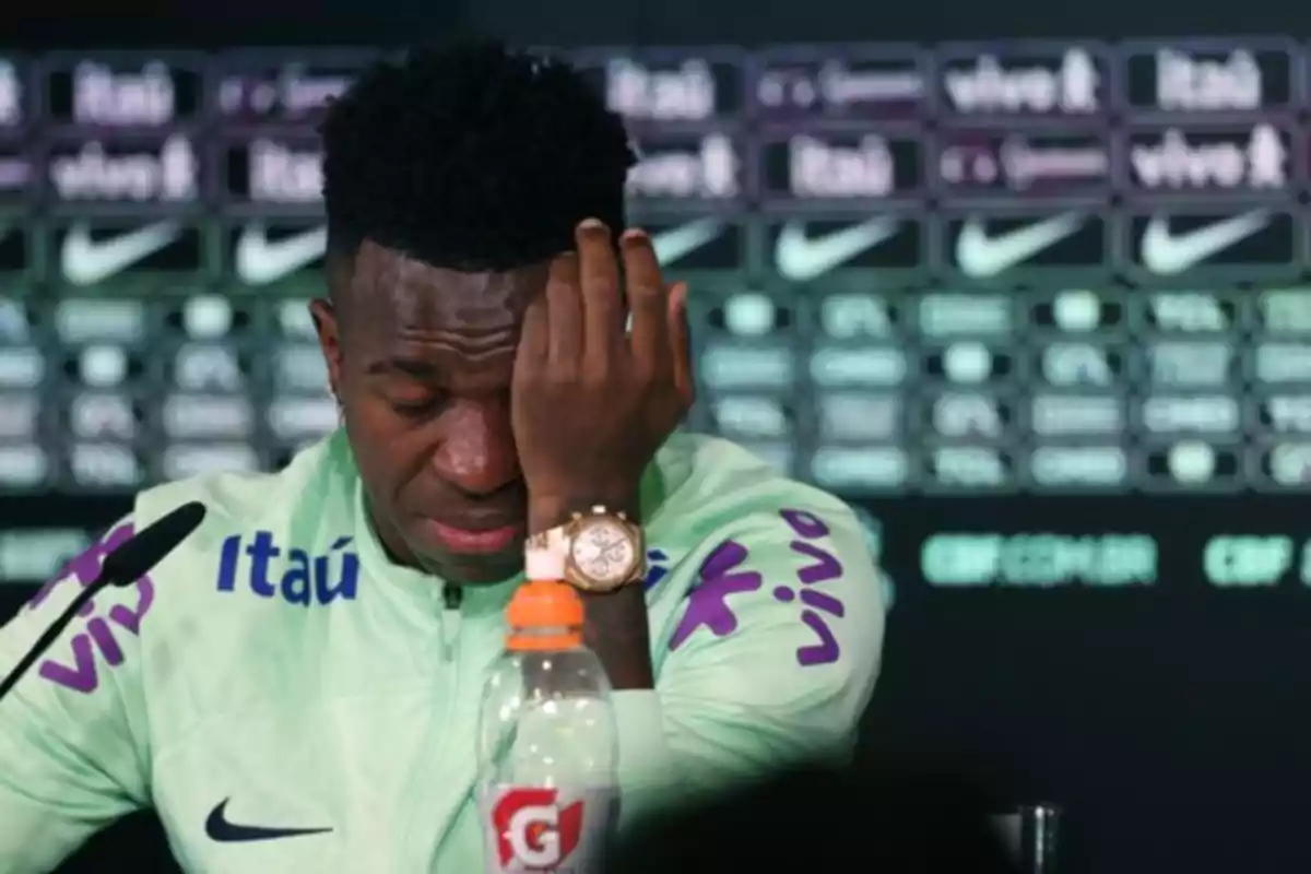 A soccer player at a press conference with a tired expression and a hand on his face.