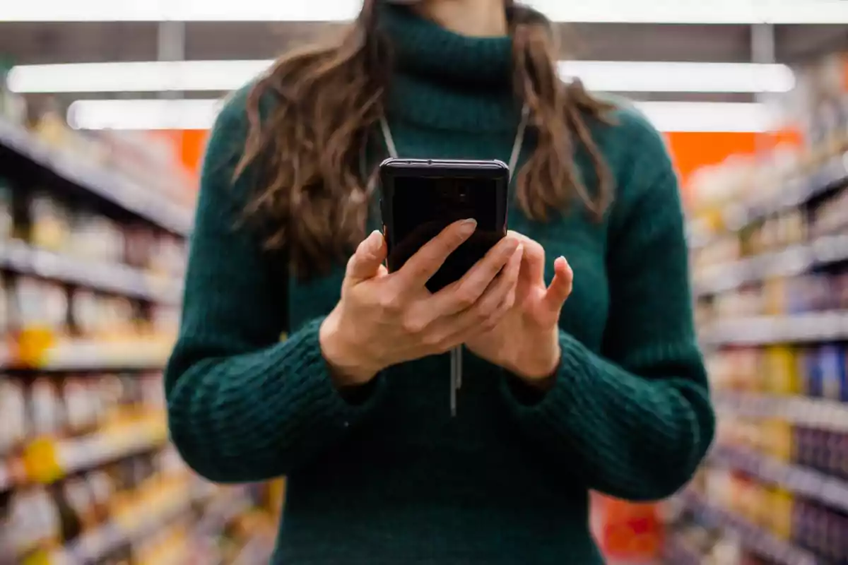 Mujer con suéter verde usando un teléfono móvil en un pasillo de supermercado Mujer con suéter verde usando un teléfono móvil en un pasillo de supermercado