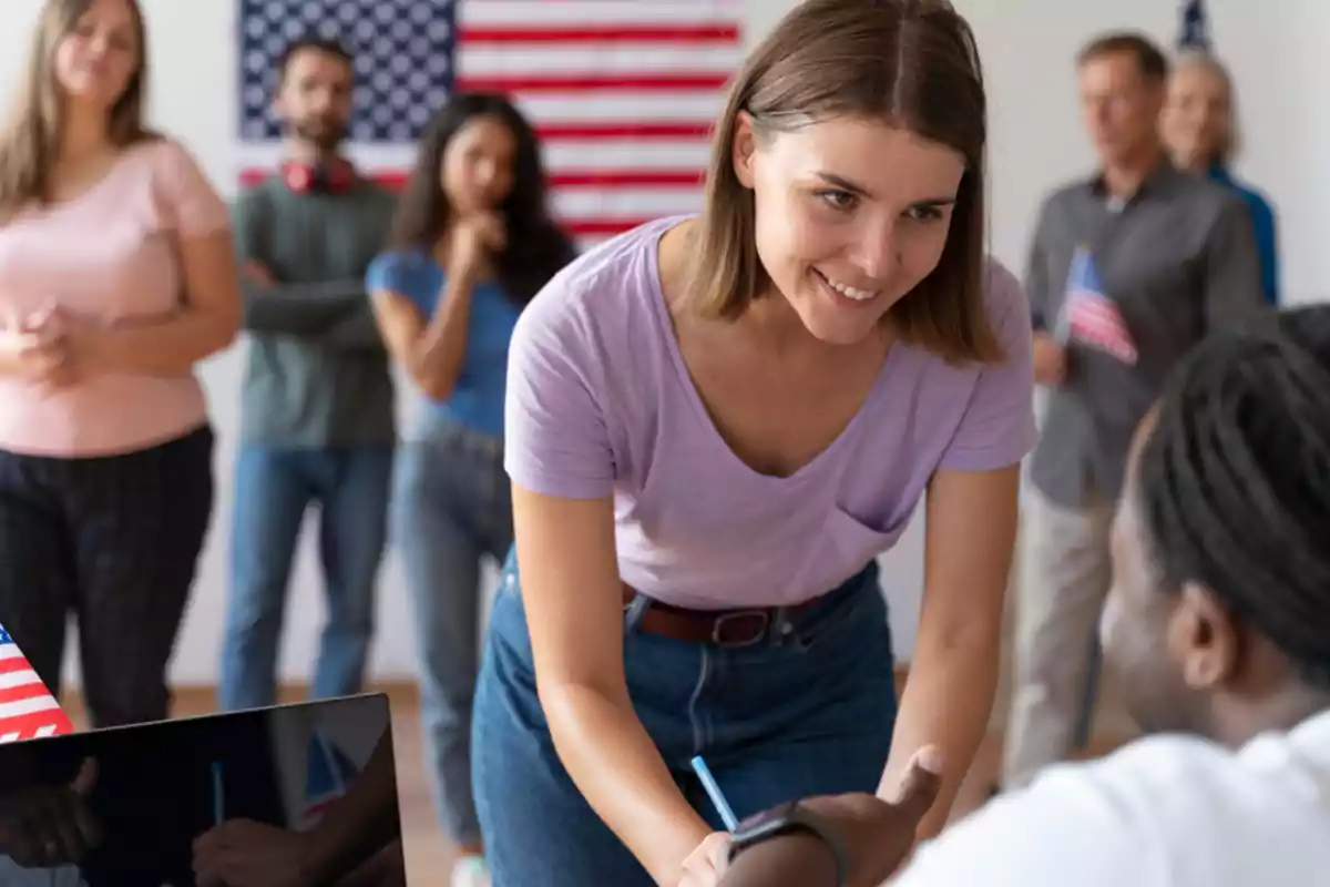 Mujer joven sonriente entregando un documento a una persona sentada mientras otras personas esperan en fila con una bandera de Estados Unidos al fondo Mujer joven sonriente entregando un documento a una persona sentada mientras otras personas esperan en fila con una bandera de Estados Unidos al fondo