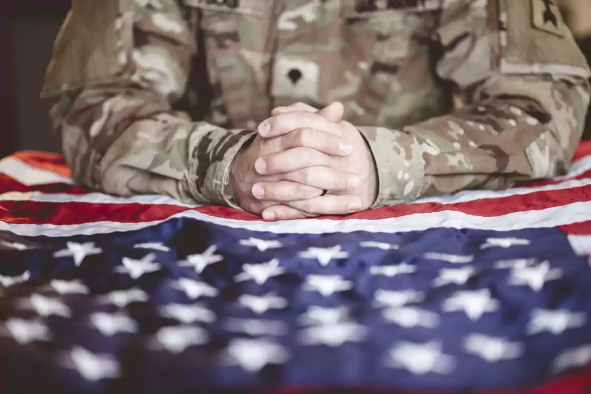 Persona con uniforme militar apoya las manos entrelazadas sobre una bandera de Estados Unidos