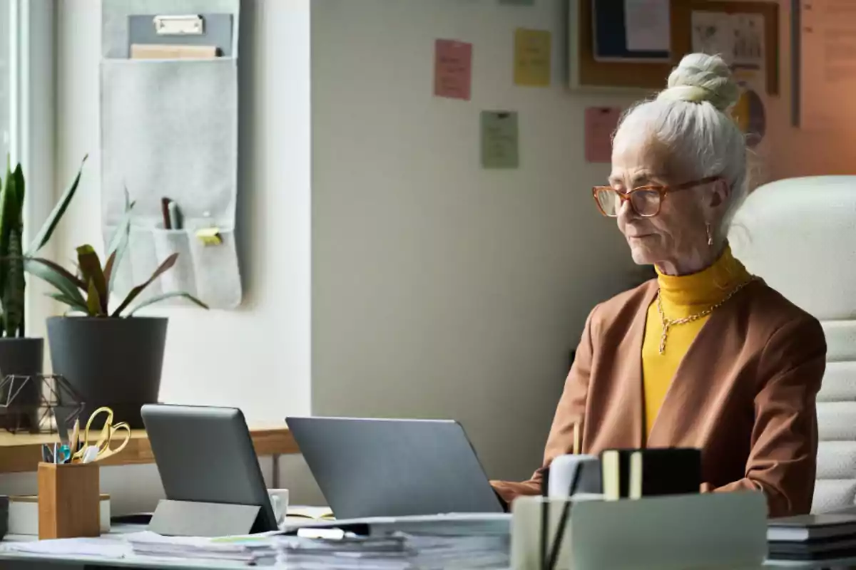 Mujer mayor con gafas y cabello recogido trabajando en una oficina moderna frente a una computadora portátil rodeada de plantas y material de oficina Mujer mayor con gafas y cabello recogido trabajando en una oficina moderna frente a una computadora portátil rodeada de plantas y material de oficina