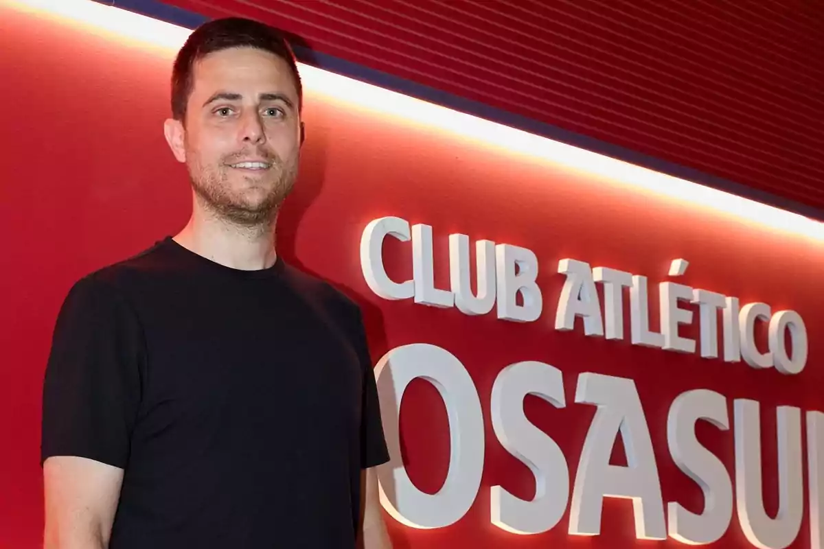 Hombre con camiseta negra posando frente a una pared roja con el letrero Club Atlético Osasuna Hombre con camiseta negra posando frente a una pared roja con el letrero Club Atlético Osasuna