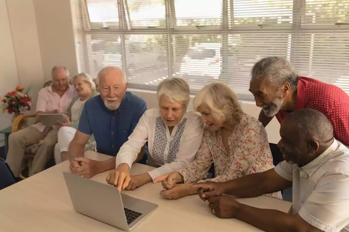 Grupo de personas mayores sentadas alrededor de una mesa usando una computadora portátil y sonriendo mientras interactúan juntos en un ambiente luminoso Grupo de personas mayores sentadas alrededor de una mesa usando una computadora portátil y sonriendo mientras interactúan juntos en un ambiente luminoso