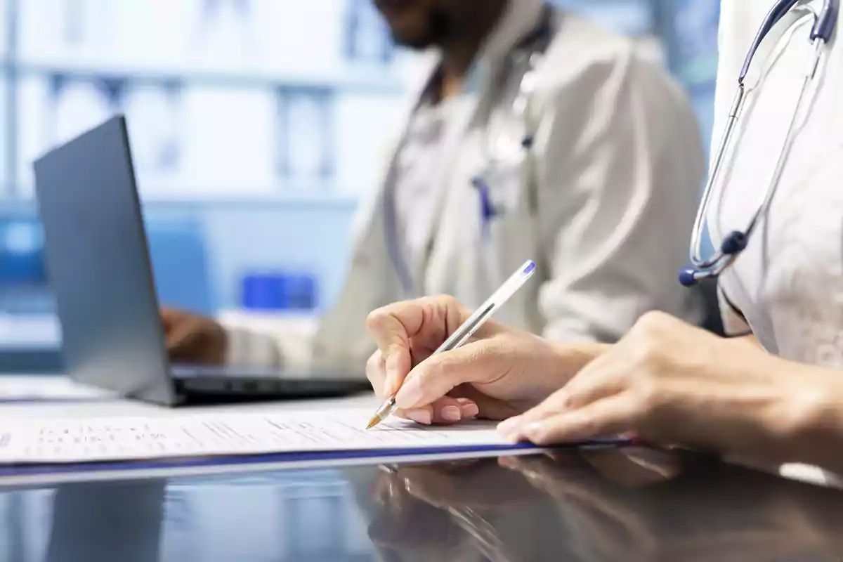 Dos profesionales de la salud con bata blanca y estetoscopio revisan documentos médicos y utilizan una computadora portátil en un entorno clínico. Dos profesionales de la salud con bata blanca y estetoscopio revisan documentos médicos y utilizan una computadora portátil en un entorno clínico.