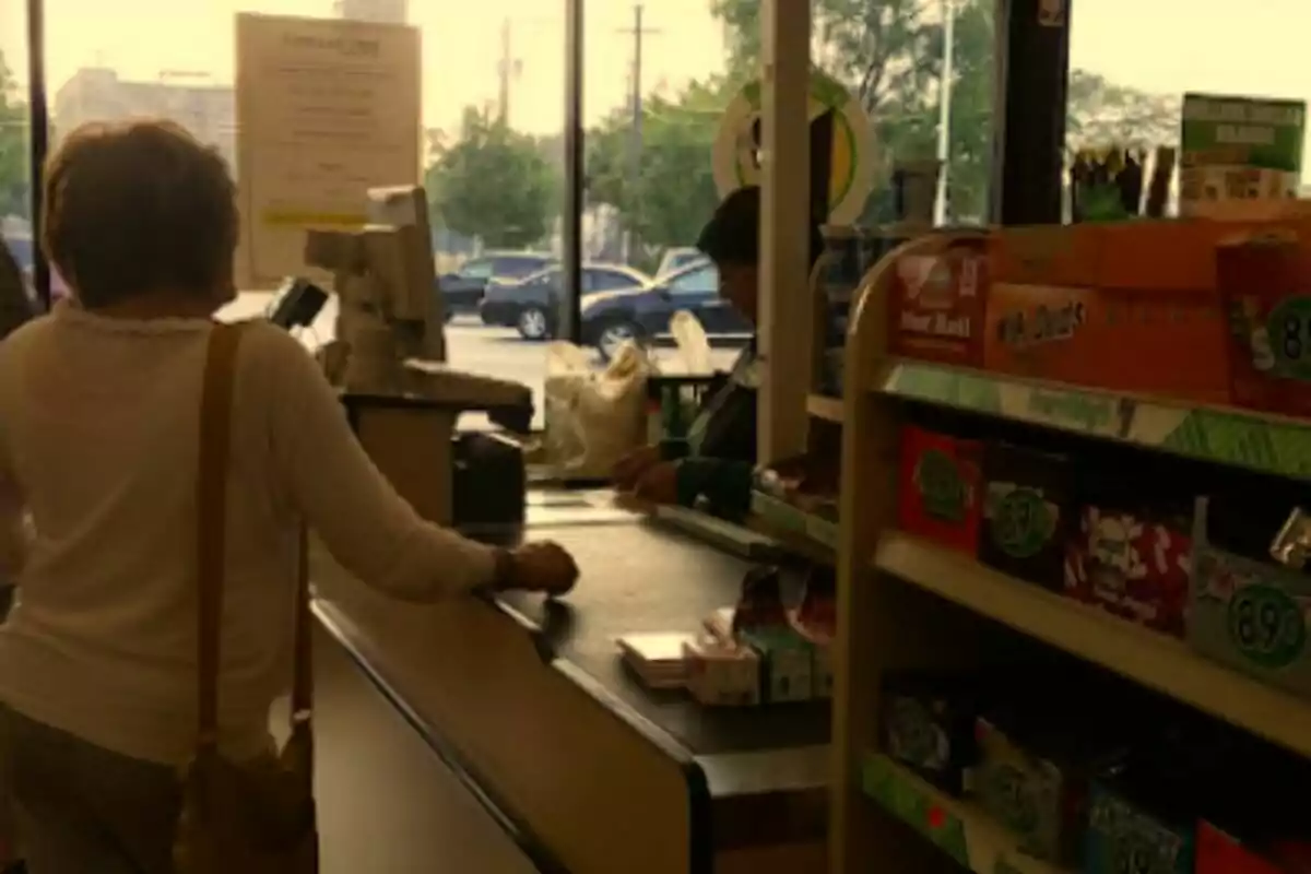 A person is paying at a store checkout while another person is working behind the counter, and there is a shelf with candy next to them.