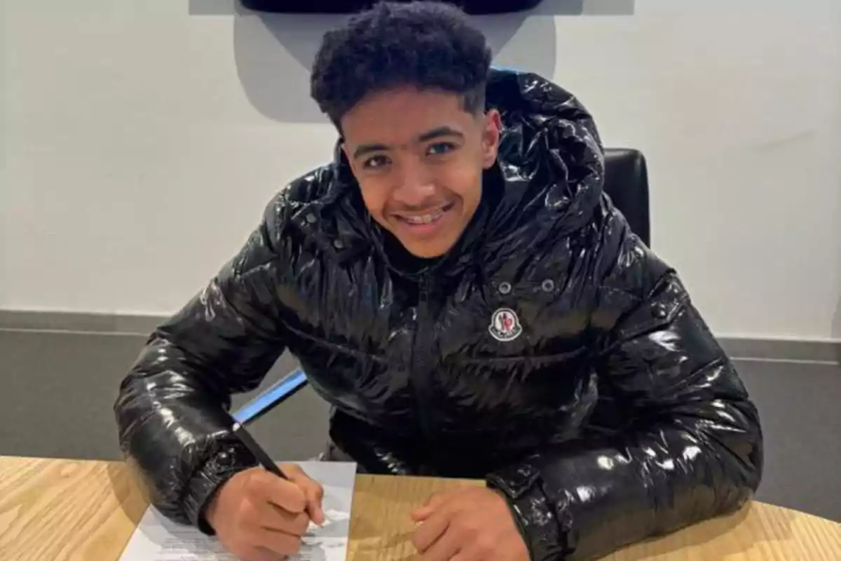Smiling young man in a shiny black jacket signing a document at a wooden table
