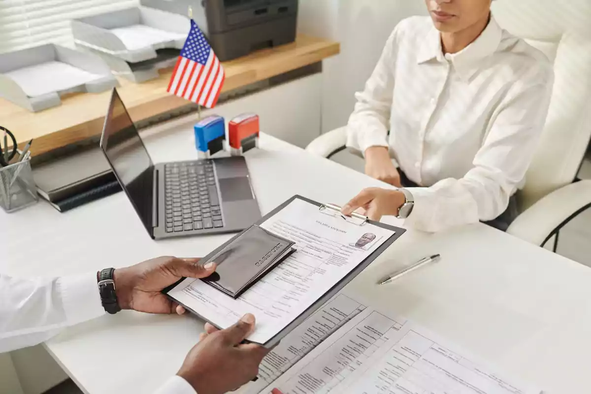 Persona entregando documentos y pasaporte a una funcionaria en una oficina con una bandera de Estados Unidos y una computadora portátil sobre el escritorio Persona entregando documentos y pasaporte a una funcionaria en una oficina con una bandera de Estados Unidos y una computadora portátil sobre el escritorio