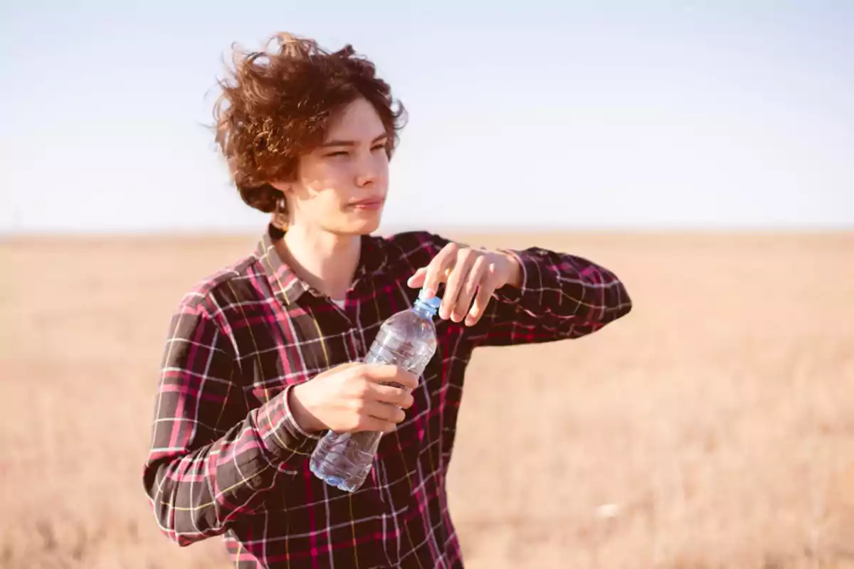 Joven con camisa de cuadros sosteniendo una botella de agua en un campo seco Joven con camisa de cuadros sosteniendo una botella de agua en un campo seco