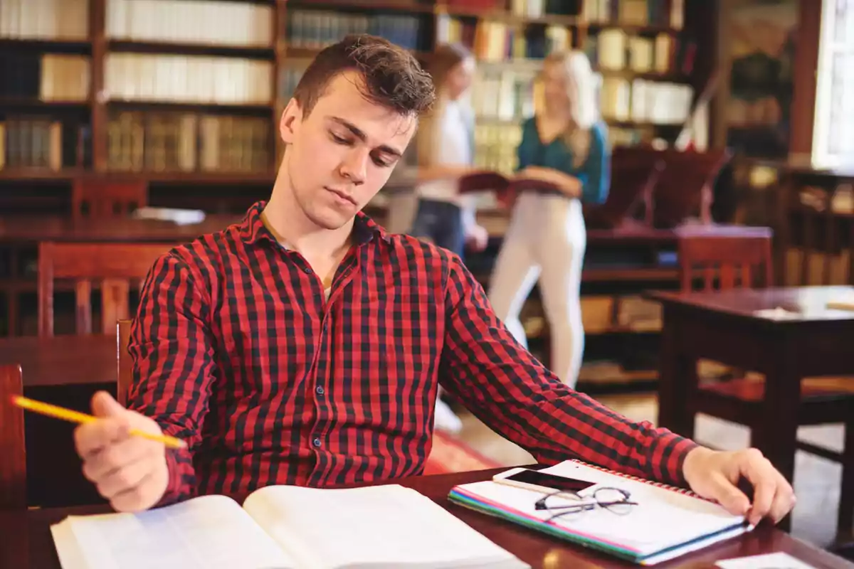 Joven con camisa de cuadros rojos estudiando en una biblioteca con libros abiertos y dos personas conversando al fondo Joven con camisa de cuadros rojos estudiando en una biblioteca con libros abiertos y dos personas conversando al fondo