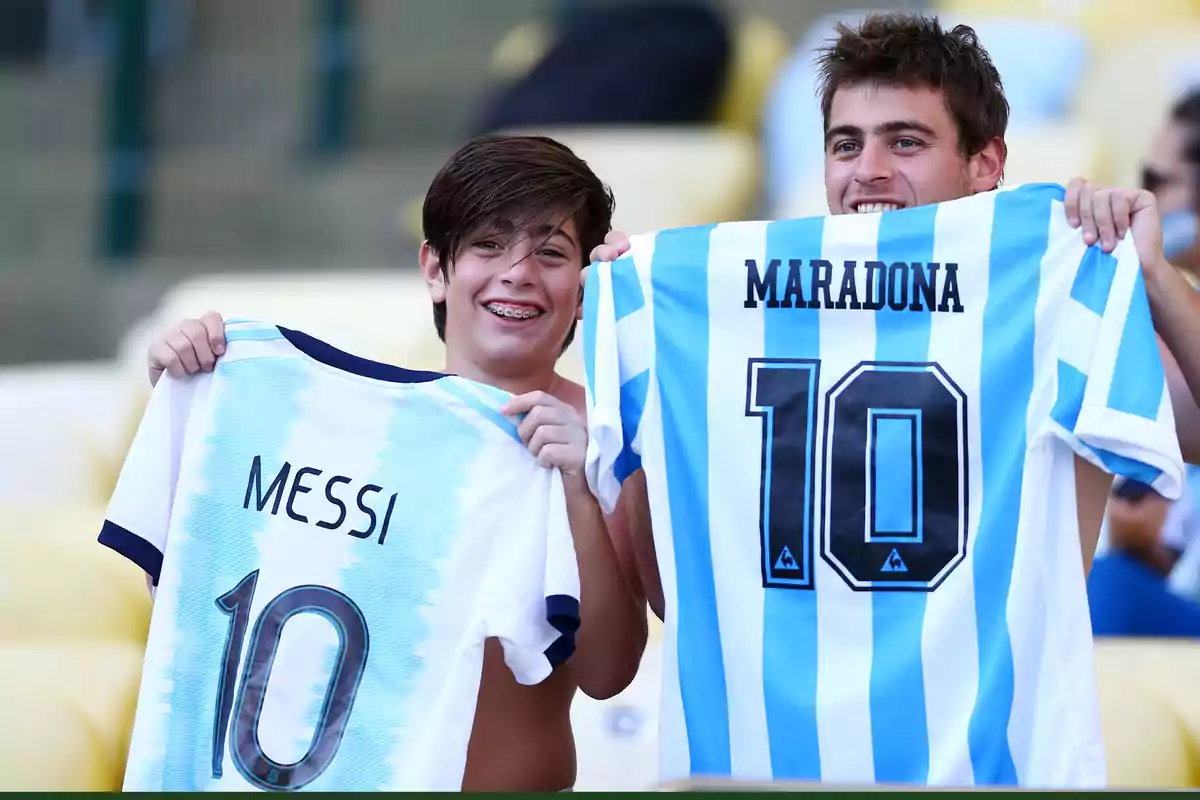 Two smiling fans hold Argentina national team jerseys, one with Messi’s name and the other with Maradona’s, both with the number 10. Two smiling fans hold Argentina national team jerseys, one with Messi’s name and the other with Maradona’s, both with the number 10.