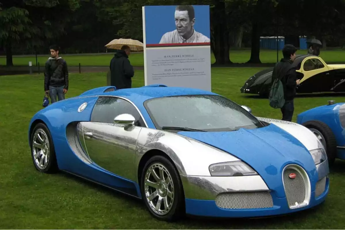 Un Bugatti Veyron azul y plateado está estacionado sobre césped mojado en una exhibición de autos, con varias personas y otros vehículos clásicos al fondo. Un Bugatti Veyron azul y plateado está estacionado sobre césped mojado en una exhibición de autos, con varias personas y otros vehículos clásicos al fondo.