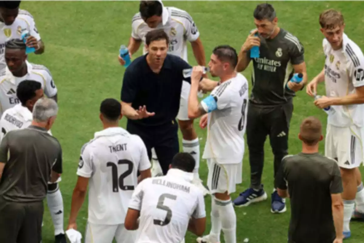 A group of players dressed in white uniforms and coaches gathered on the field during a break, some drinking water while listening to instructions.