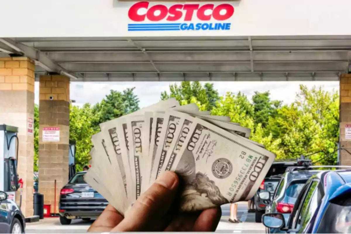 Una mano sostiene varios billetes de cien dólares frente a una gasolinera de Costco con autos haciendo fila