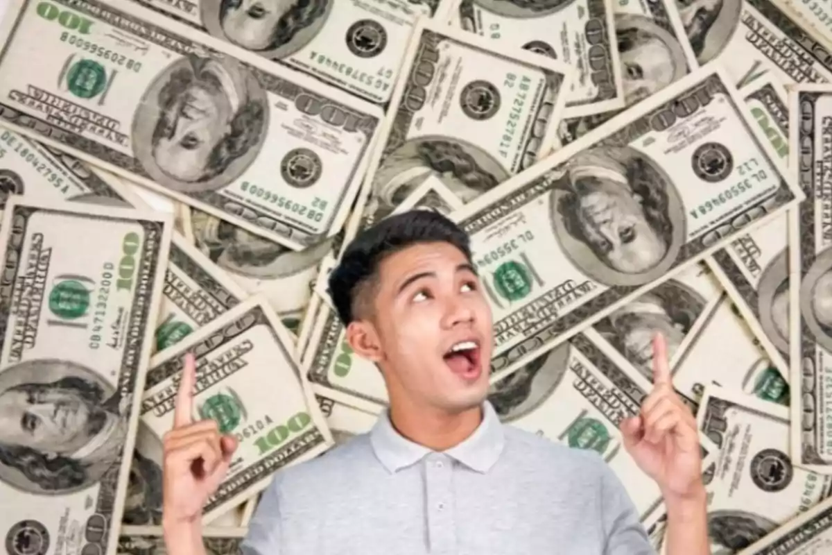 Hombre joven sonriente señalando hacia arriba con ambas manos frente a un fondo de billetes de cien dólares estadounidenses Hombre joven sonriente señalando hacia arriba con ambas manos frente a un fondo de billetes de cien dólares estadounidenses