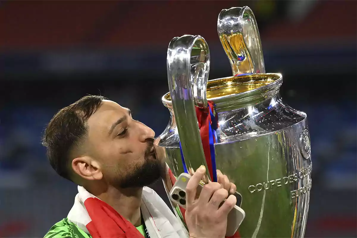 Futbolista besando un trofeo grande de plata mientras sostiene un teléfono móvil y lleva una bandera sobre los hombros