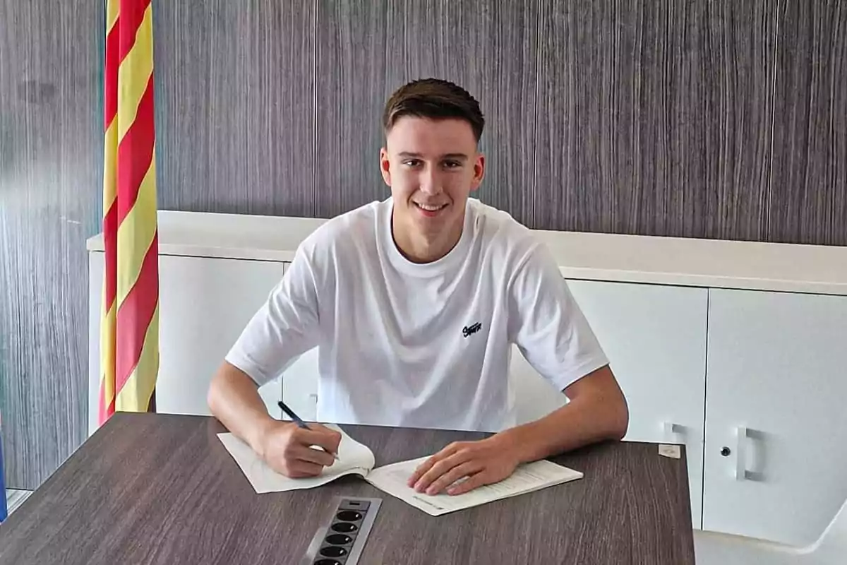 Joven sonriente con camiseta blanca firmando un documento en una mesa de oficina junto a una bandera de rayas rojas y amarillas Joven sonriente con camiseta blanca firmando un documento en una mesa de oficina junto a una bandera de rayas rojas y amarillas