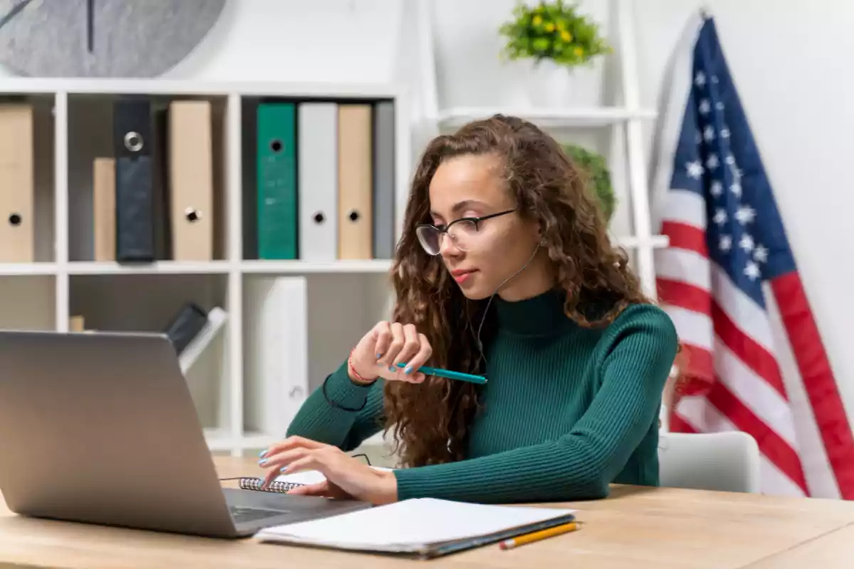 Joven con gafas y suéter verde trabajando en una laptop en una oficina con estantería y bandera de Estados Unidos al fondo Joven con gafas y suéter verde trabajando en una laptop en una oficina con estantería y bandera de Estados Unidos al fondo