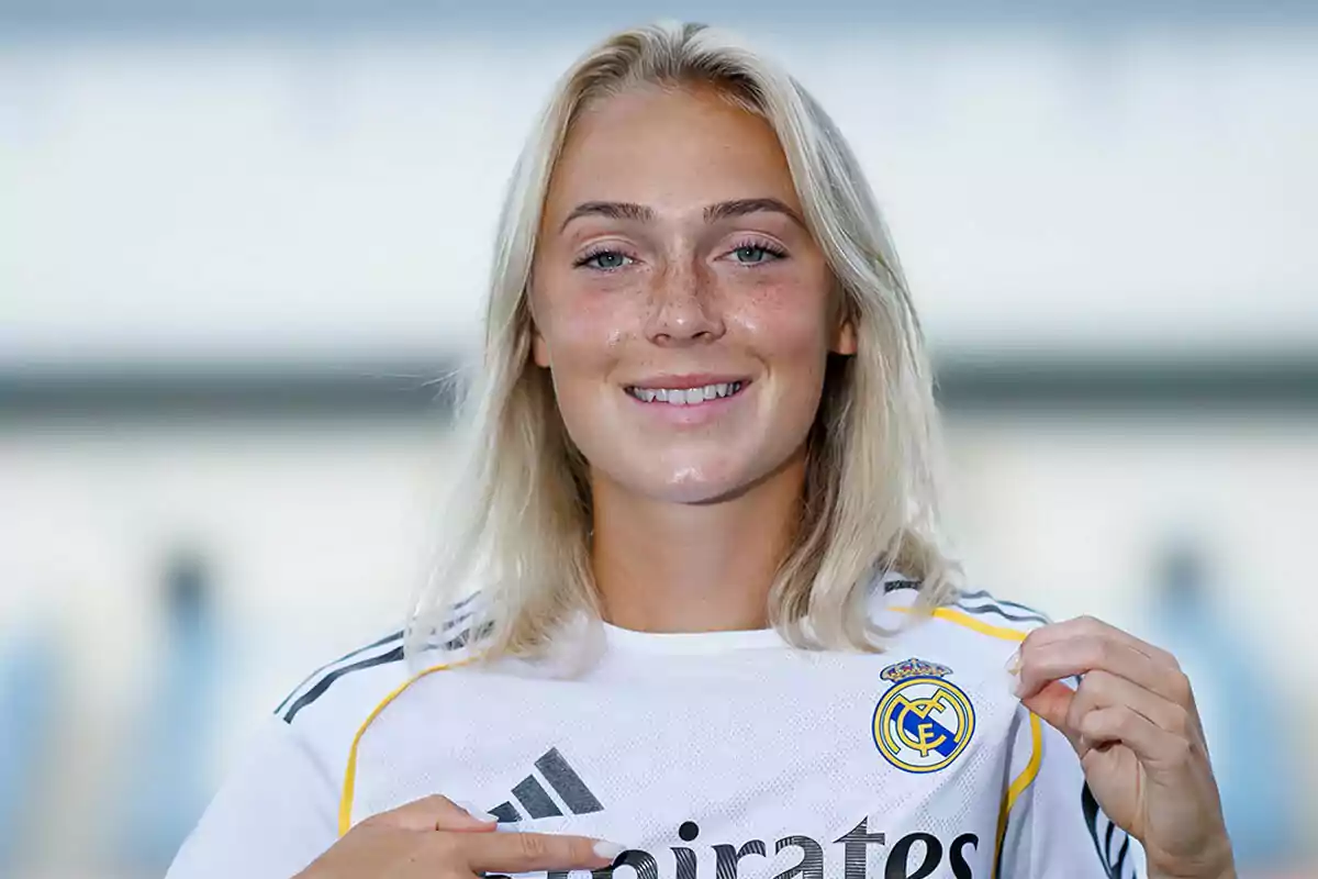 Mujer rubia sonriente con camiseta blanca del Real Madrid señalando el escudo del equipo Mujer rubia sonriente con camiseta blanca del Real Madrid señalando el escudo del equipo