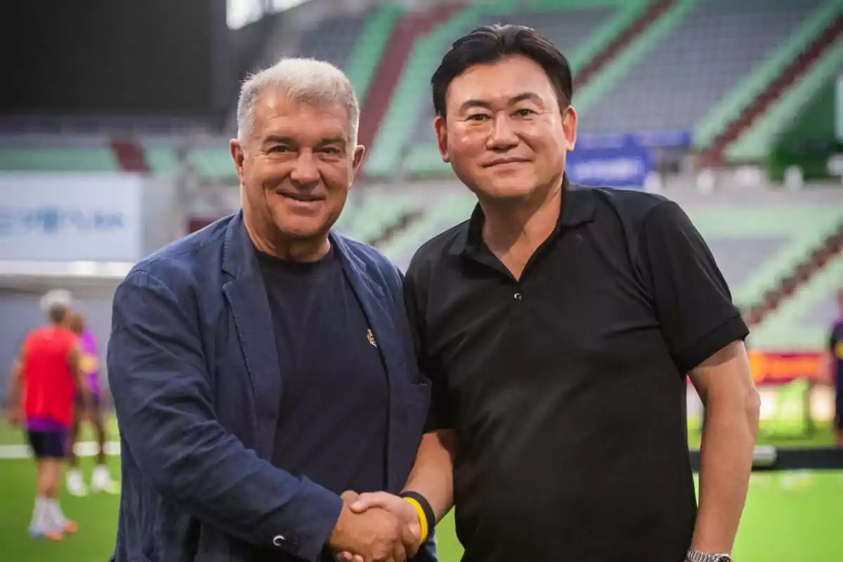 Dos hombres sonríen y se dan la mano en un estadio de fútbol con gradas verdes y rojas de fondo Dos hombres sonríen y se dan la mano en un estadio de fútbol con gradas verdes y rojas de fondo