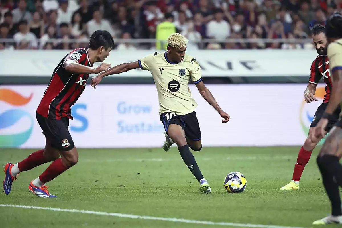Un jugador de fútbol con uniforme amarillo y negro patea el balón mientras es defendido por rivales con uniforme rojo y negro en un estadio lleno de espectadores. Un jugador de fútbol con uniforme amarillo y negro patea el balón mientras es defendido por rivales con uniforme rojo y negro en un estadio lleno de espectadores.