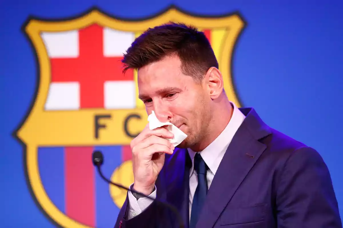 Man in a suit wiping his tears with a handkerchief in front of a microphone and a soccer club crest in the background