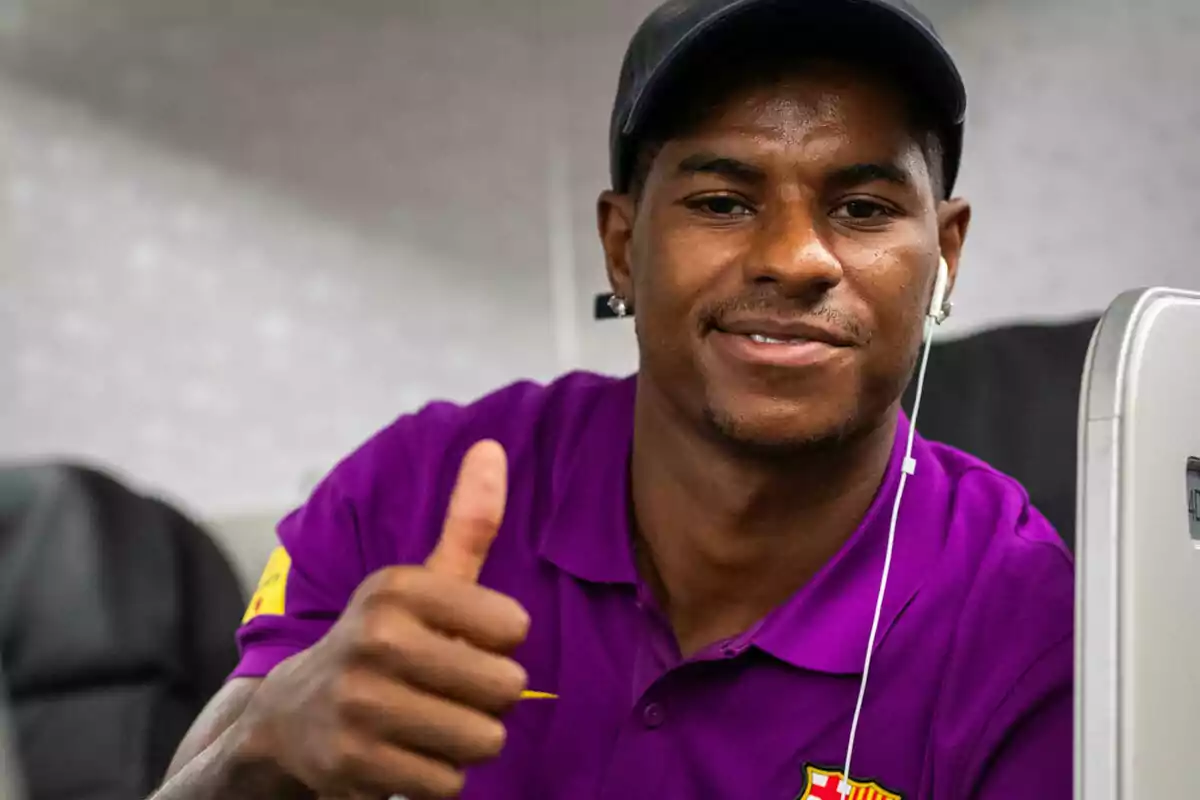 Hombre joven con camisa morada y gorra negra sonríe y muestra un pulgar arriba mientras escucha música con auriculares.