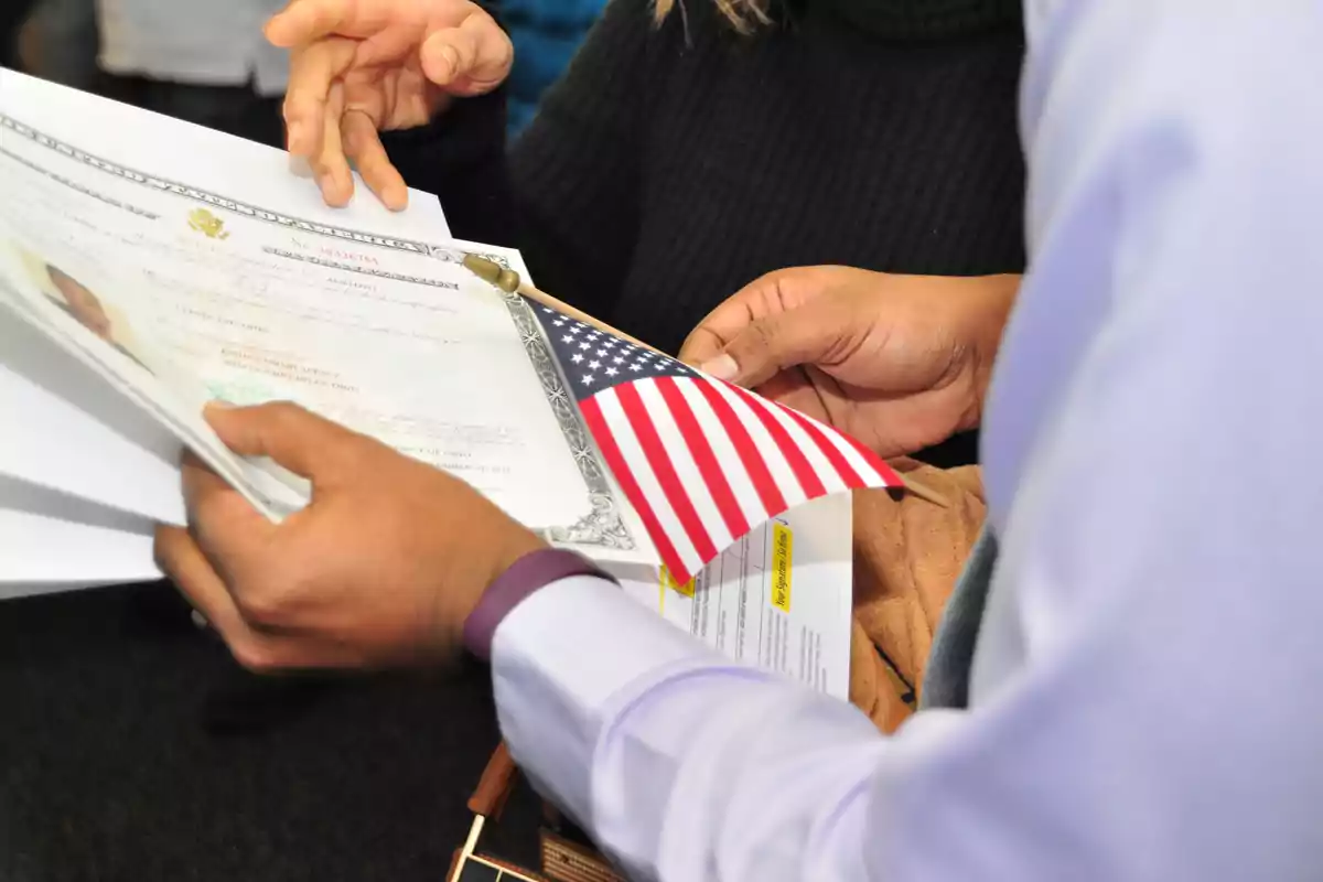 Persona sosteniendo documentos oficiales y una pequeña bandera de Estados Unidos mientras otra persona señala con la mano Persona sosteniendo documentos oficiales y una pequeña bandera de Estados Unidos mientras otra persona señala con la mano