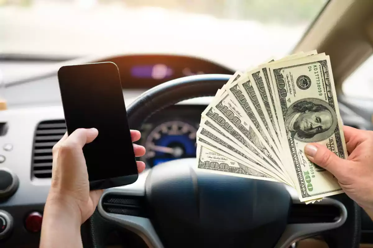 Person inside a car holding a mobile phone in one hand and several hundred-dollar bills in the other in front of the steering wheel