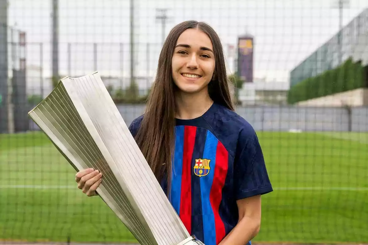 Joven con camiseta del FC Barcelona sonríe mientras sostiene un gran trofeo en un campo de fútbol Joven con camiseta del FC Barcelona sonríe mientras sostiene un gran trofeo en un campo de fútbol