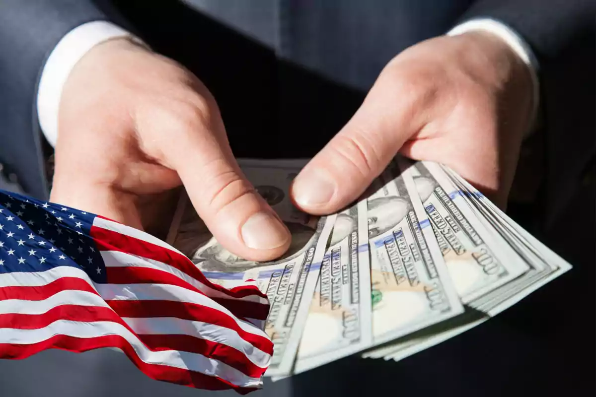 Manos sosteniendo billetes de cien dólares junto a una bandera de Estados Unidos