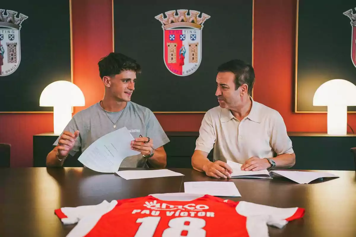 Dos personas sentadas en una mesa firmando documentos con una camiseta de fútbol roja y blanca sobre la mesa y el escudo del club Braga en la pared de fondo