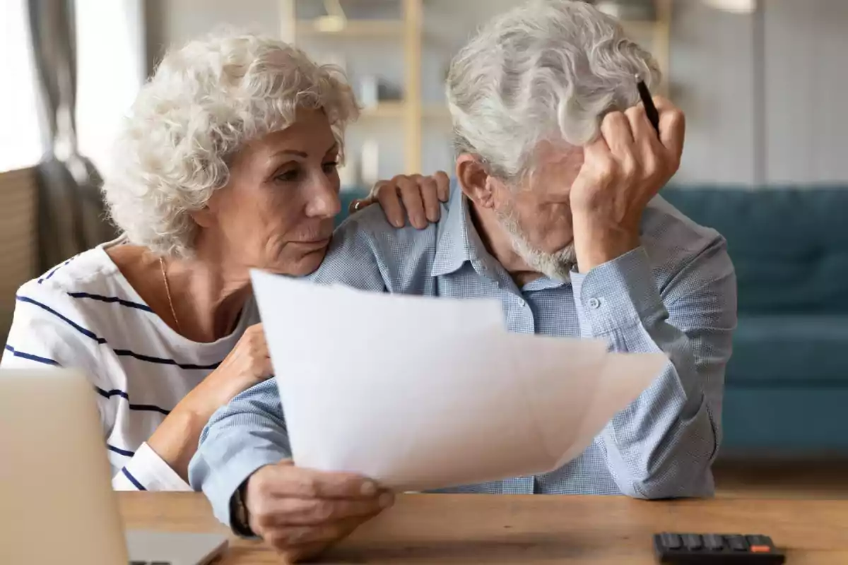 Pareja de personas mayores sentadas en una mesa revisando documentos con expresión preocupada Pareja de personas mayores sentadas en una mesa revisando documentos con expresión preocupada