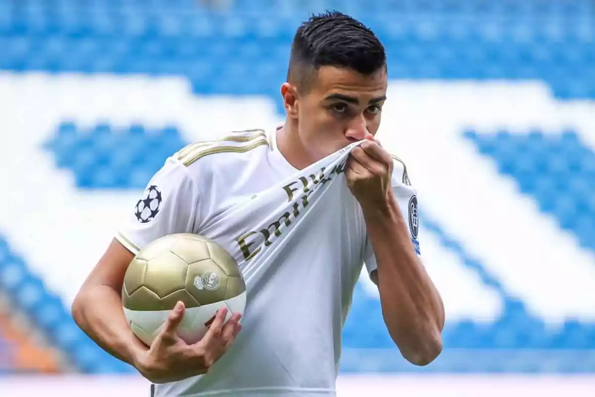 Soccer player in a white uniform kissing the crest on his jersey and holding a golden ball in a stadium Soccer player in a white uniform kissing the crest on his jersey and holding a golden ball in a stadium