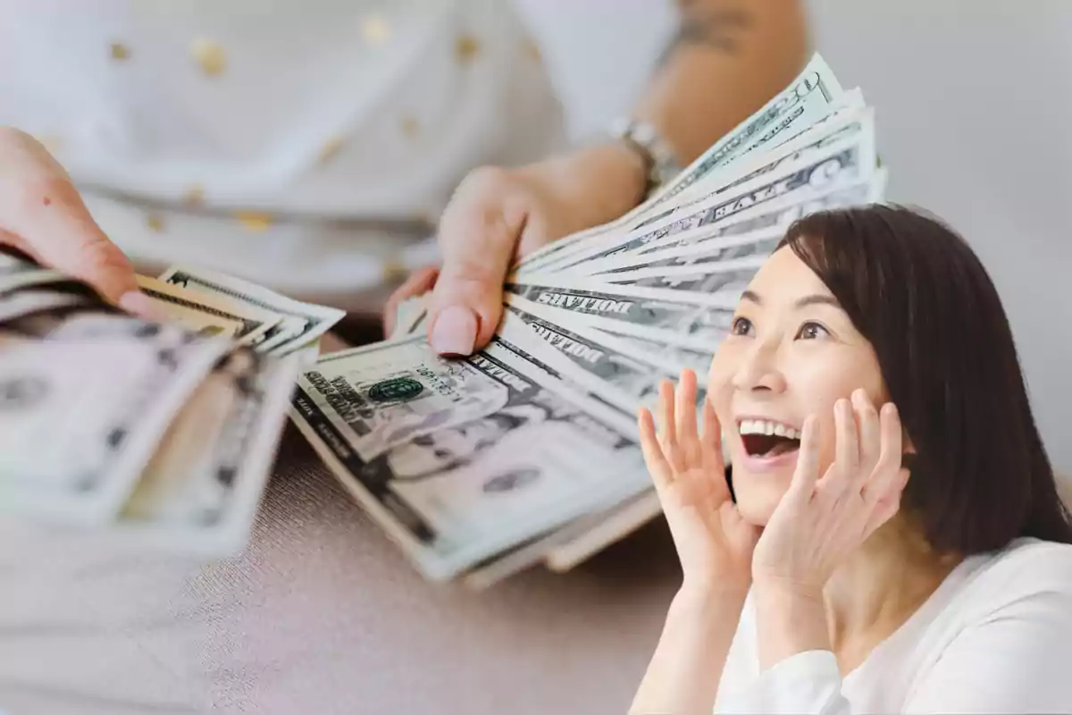 Una mujer sonriente y sorprendida observa un fajo de billetes de dólar sostenidos por otra persona. Una mujer sonriente y sorprendida observa un fajo de billetes de dólar sostenidos por otra persona.