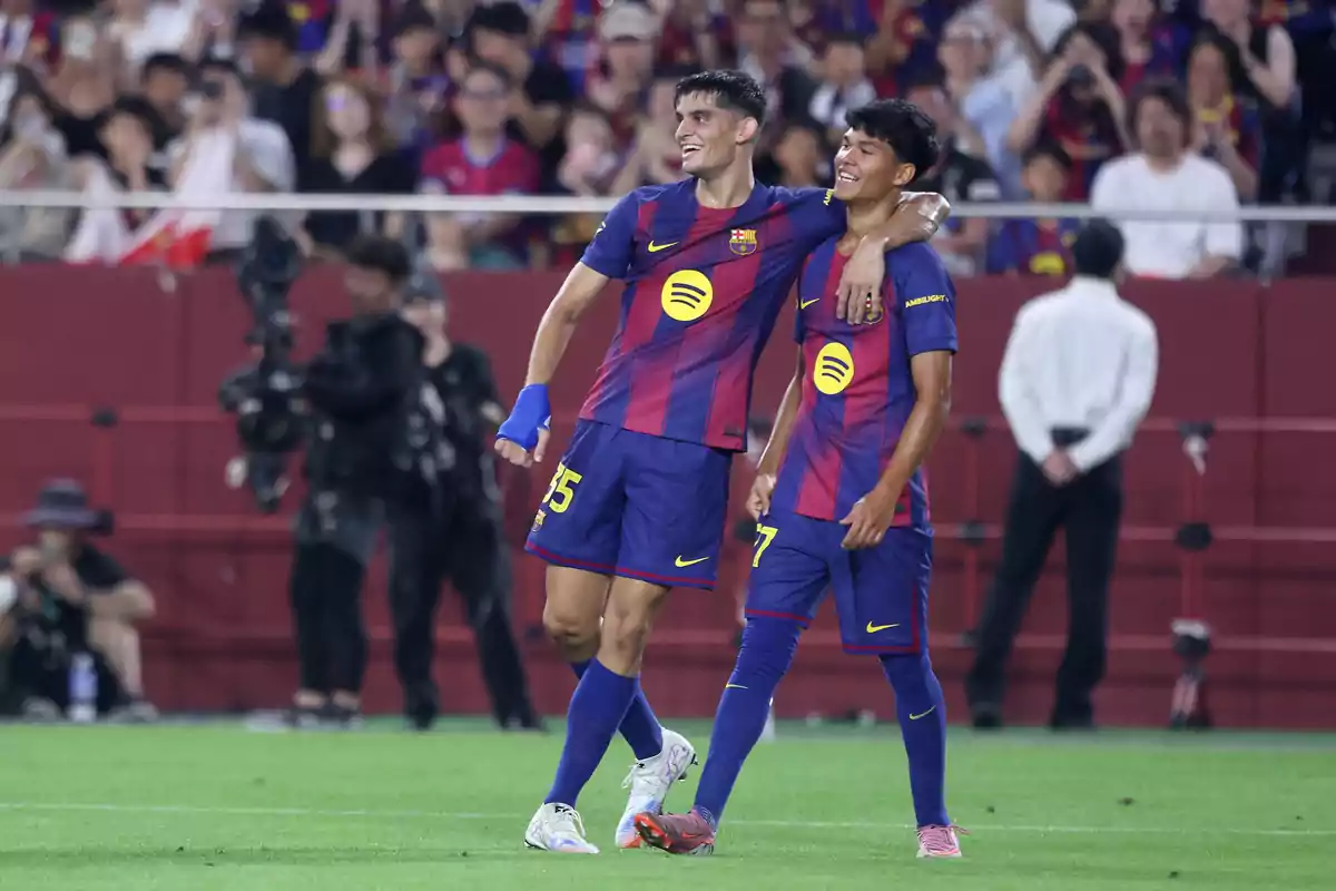 Dos jugadores del Barcelona sonríen y se abrazan en el campo durante un partido mientras el público los observa desde las gradas Dos jugadores del Barcelona sonríen y se abrazan en el campo durante un partido mientras el público los observa desde las gradas