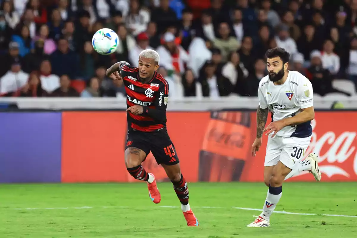 Two players compete for the ball during a match, one wearing a red and black uniform and the other in white, while the crowd watches from the stands. Two players compete for the ball during a match, one wearing a red and black uniform and the other in white, while the crowd watches from the stands.