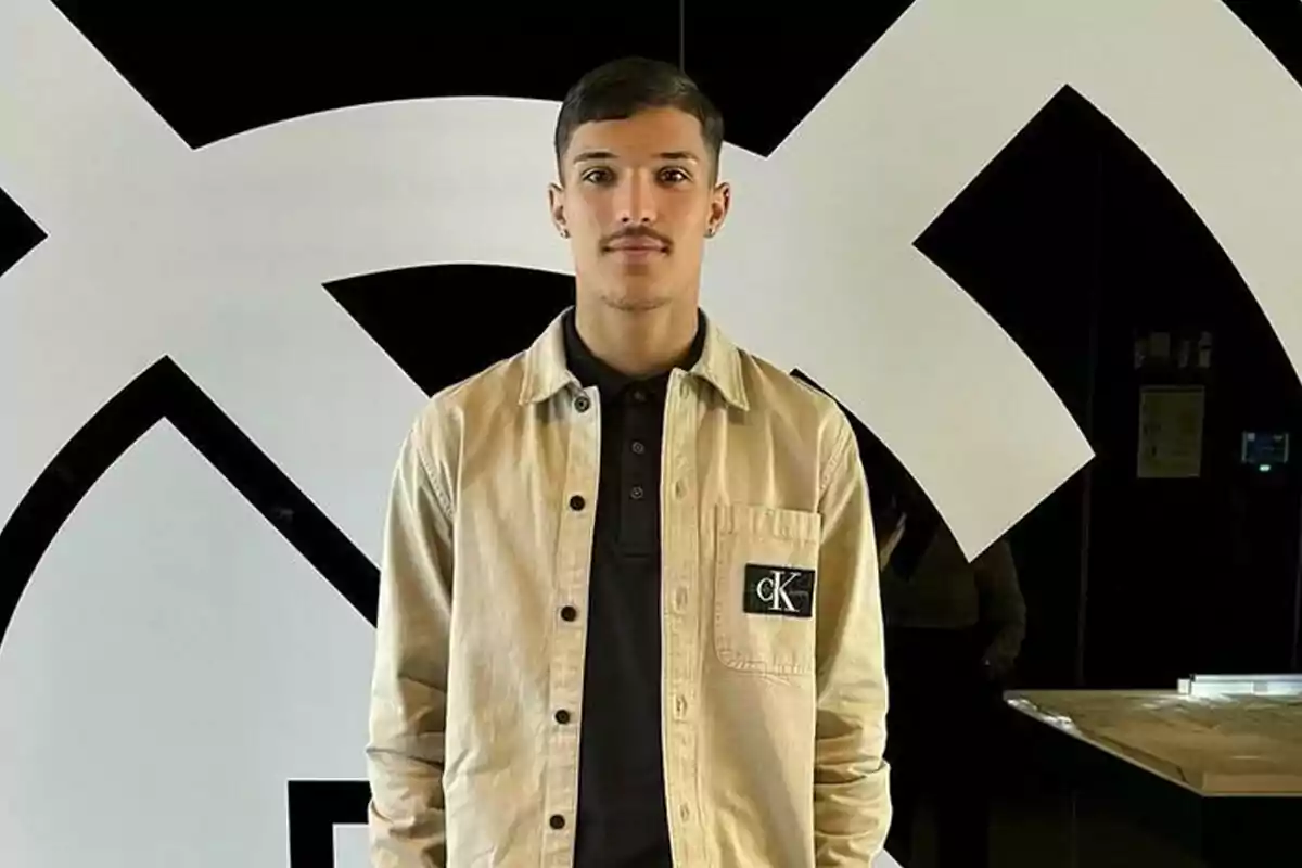 Joven de cabello corto y bigote posando frente a una pared con un diseño geométrico en blanco y negro, lleva una chaqueta beige con una camisa negra Joven de cabello corto y bigote posando frente a una pared con un diseño geométrico en blanco y negro, lleva una chaqueta beige con una camisa negra