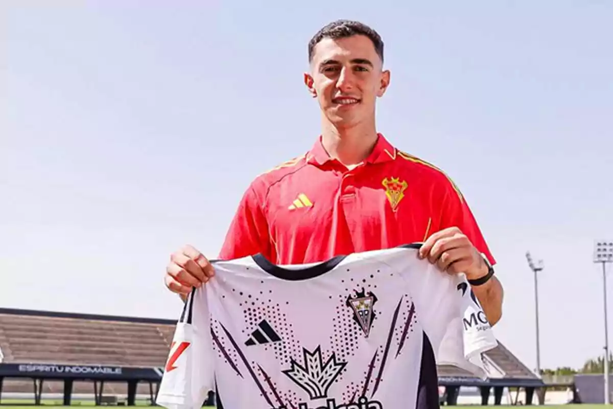 Joven con camiseta roja sostiene una camiseta de fútbol blanca en un estadio al aire libre Joven con camiseta roja sostiene una camiseta de fútbol blanca en un estadio al aire libre