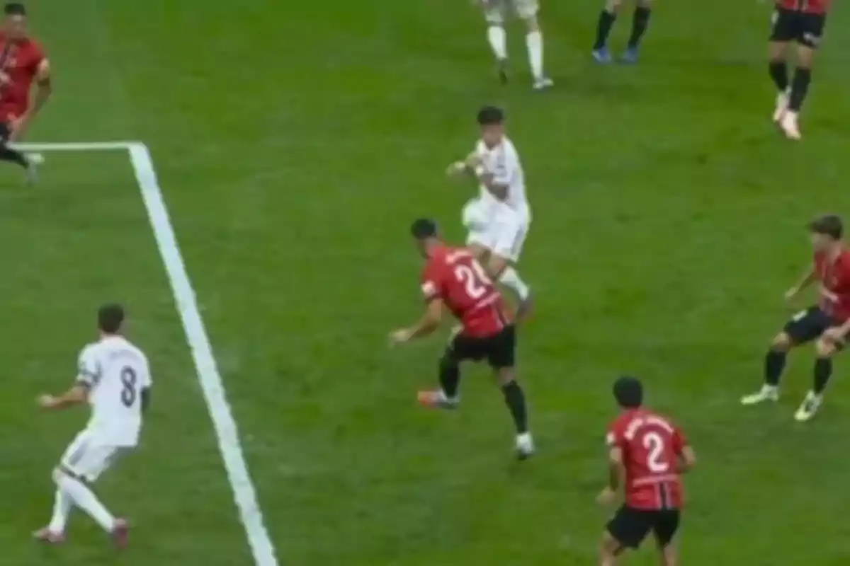 Soccer players contest the ball inside the box during a match, some wearing white uniforms and others wearing red and black uniforms.