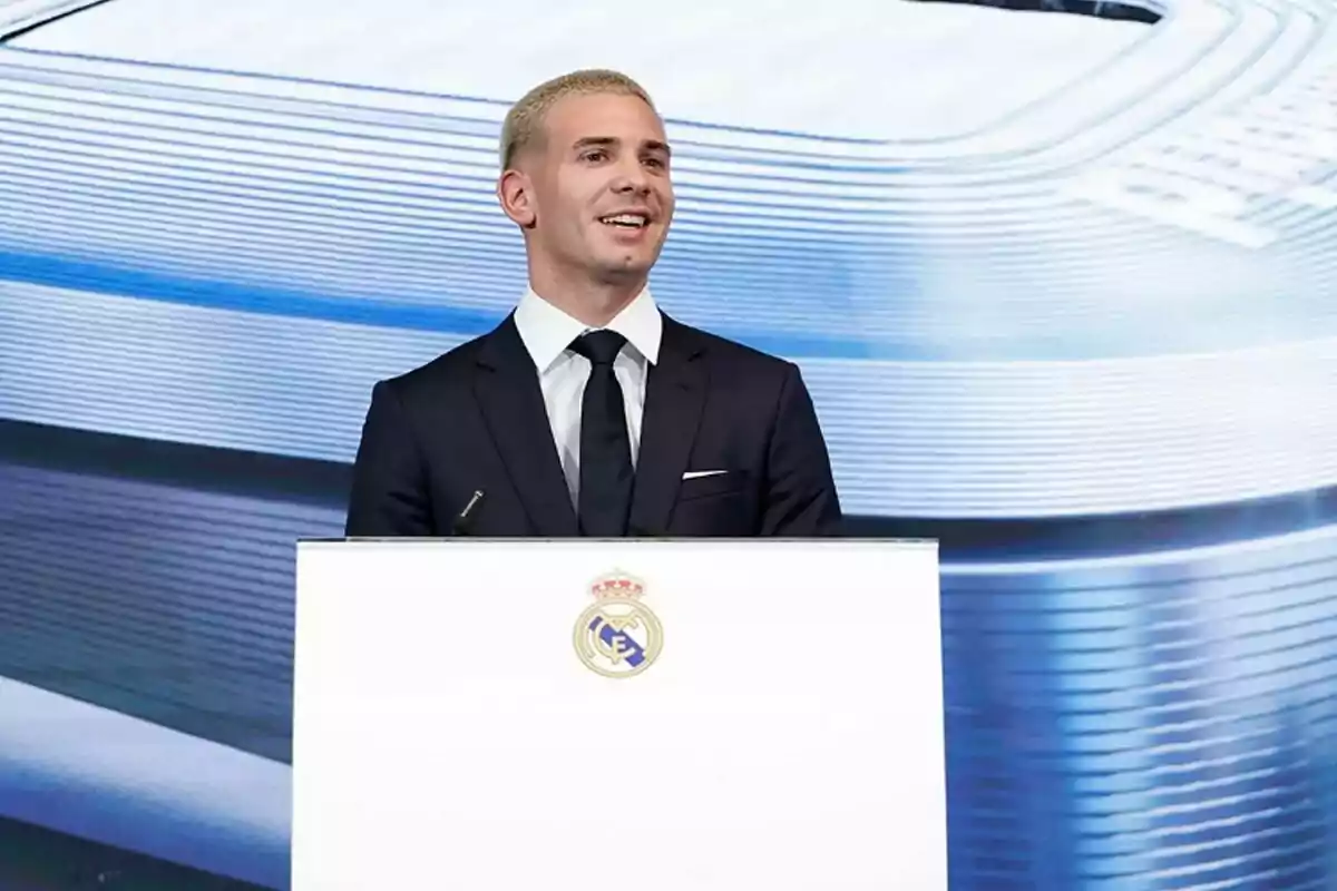 Hombre de cabello corto y rubio vestido con traje oscuro hablando en un podio con el escudo del Real Madrid al frente y un fondo azul claro. Hombre de cabello corto y rubio vestido con traje oscuro hablando en un podio con el escudo del Real Madrid al frente y un fondo azul claro.