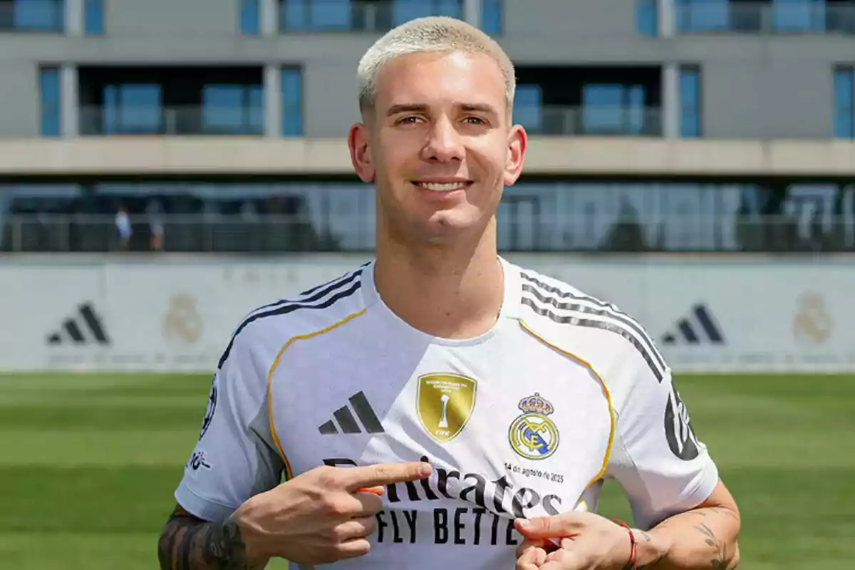 Jugador de fútbol con cabello corto y rubio sonríe mientras señala el escudo del Real Madrid en su camiseta blanca durante el día en un campo de entrenamiento