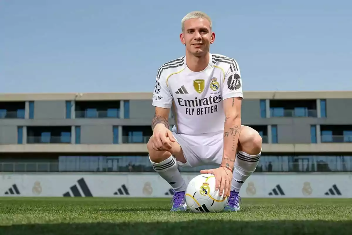 Futbolista con uniforme blanco del Real Madrid posando en una cancha de césped mientras sostiene un balón con una mano y al fondo se observa un edificio moderno Futbolista con uniforme blanco del Real Madrid posando en una cancha de césped mientras sostiene un balón con una mano y al fondo se observa un edificio moderno