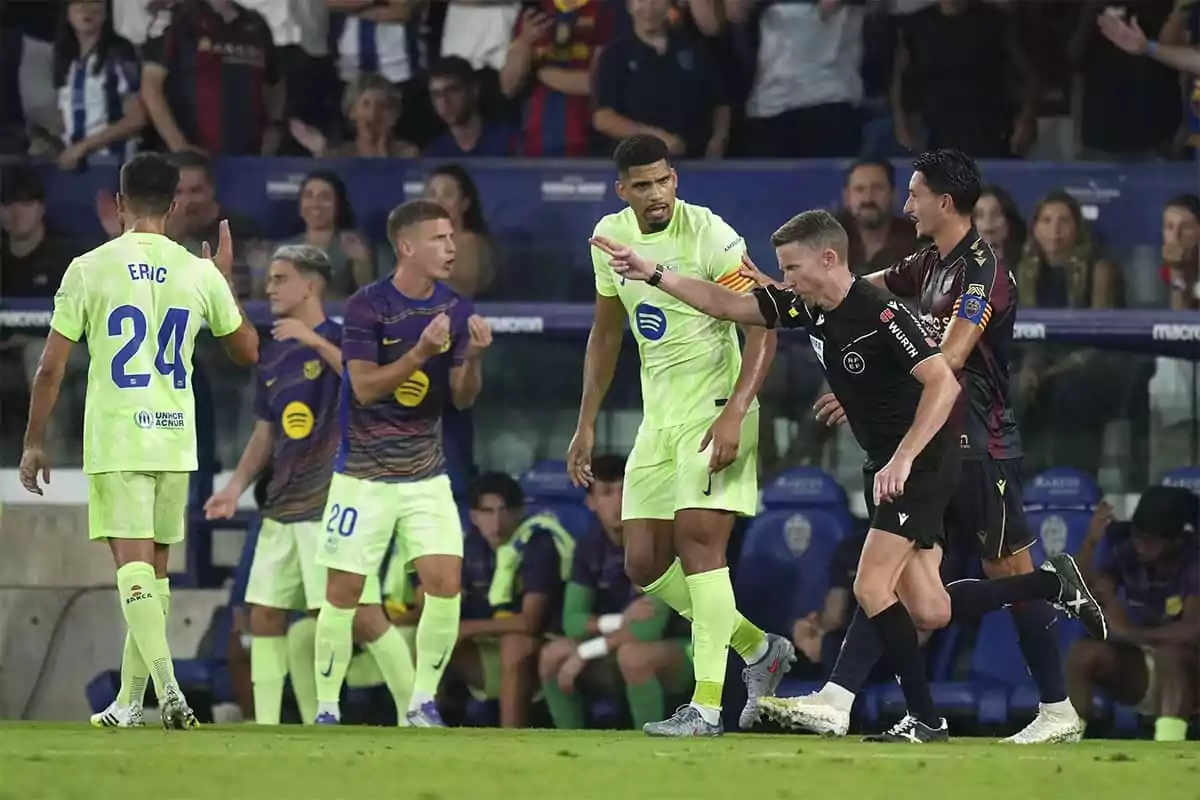 Un árbitro señala una decisión durante un partido de fútbol mientras los jugadores de ambos equipos reaccionan y el público observa desde las gradas Un árbitro señala una decisión durante un partido de fútbol mientras los jugadores de ambos equipos reaccionan y el público observa desde las gradas
