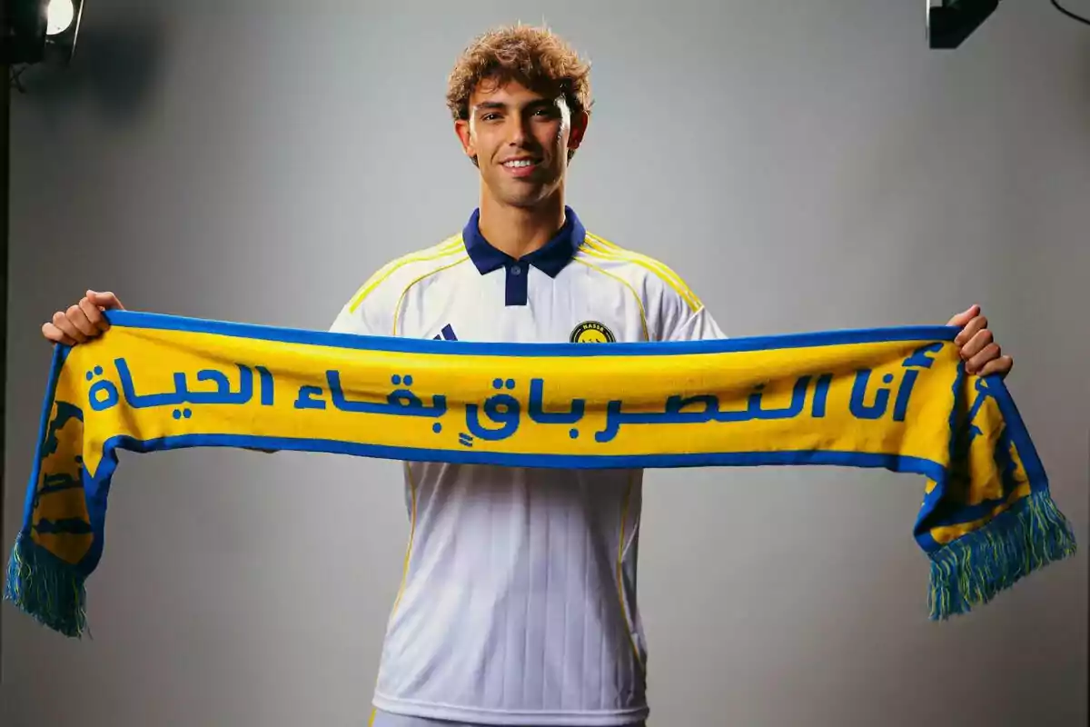 Joven con camiseta blanca de fútbol sostiene una bufanda amarilla y azul con texto en árabe, posando y sonriendo frente a la cámara en un estudio. Joven con camiseta blanca de fútbol sostiene una bufanda amarilla y azul con texto en árabe, posando y sonriendo frente a la cámara en un estudio.