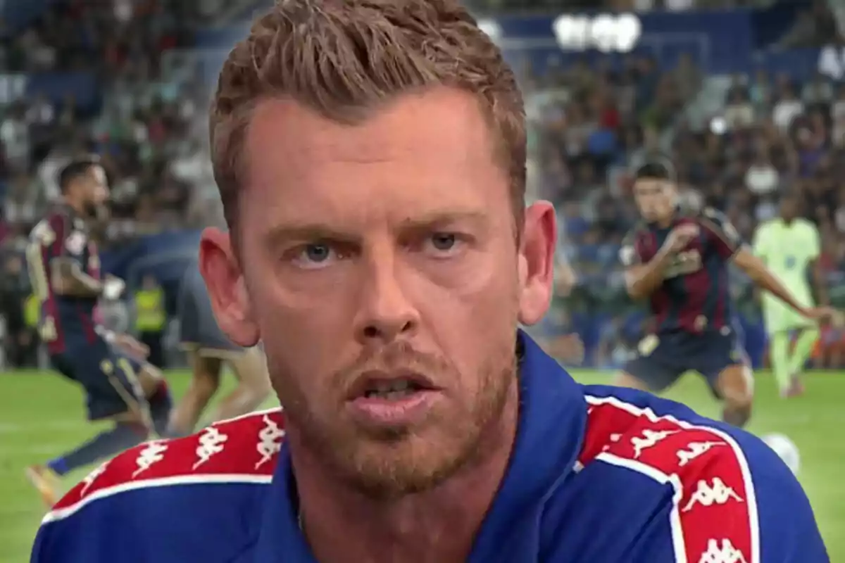 Man with a serious expression wearing a blue jacket with red and white details in a soccer stadium with players and crowd in the background