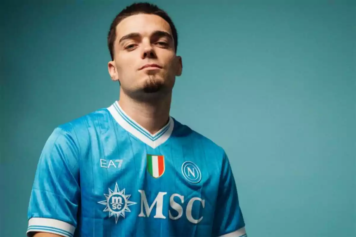 Joven con barba corta y cabello oscuro lleva puesta una camiseta azul del Napoli con fondo azul claro Joven con barba corta y cabello oscuro lleva puesta una camiseta azul del Napoli con fondo azul claro