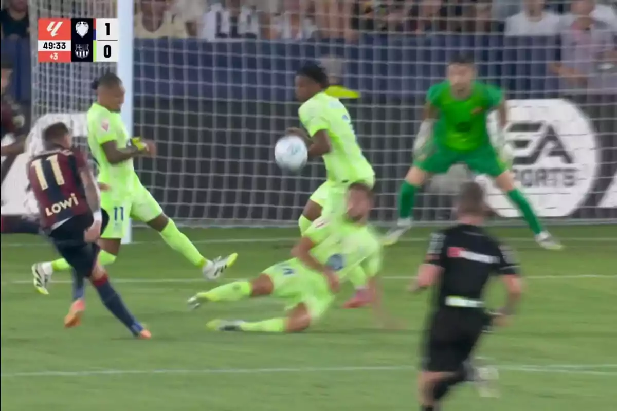 A player in a dark uniform shoots at the goal while several defenders in fluorescent uniforms try to block the ball in front of the goal, and the goalkeeper is watching the play closely.