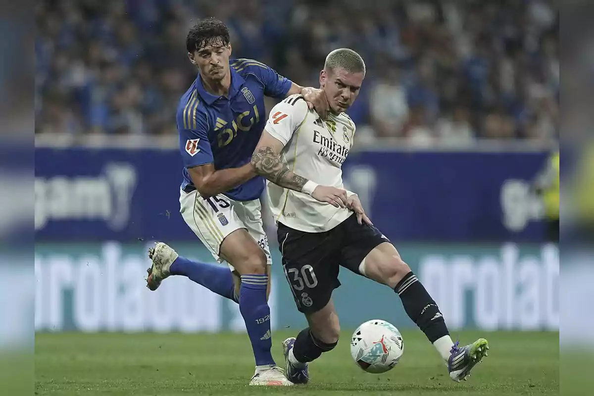 Dos futbolistas disputan el balón durante un partido, uno con uniforme azul y el otro con uniforme blanco y negro, en un estadio lleno de espectadores. Dos futbolistas disputan el balón durante un partido, uno con uniforme azul y el otro con uniforme blanco y negro, en un estadio lleno de espectadores.