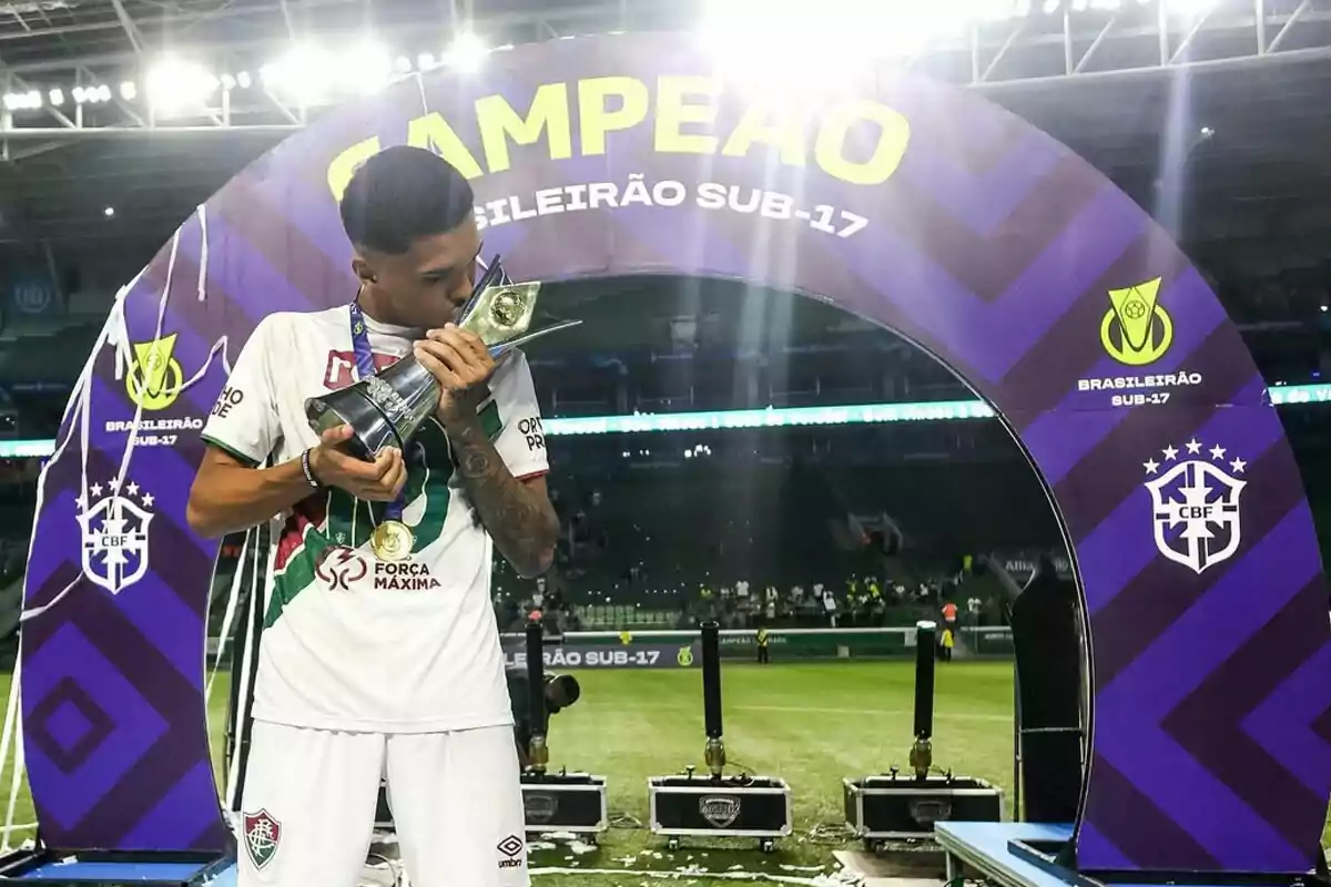 Jugador de fútbol juvenil besando un trofeo de campeón en un estadio, con un arco decorativo de fondo que celebra el título del Brasileirão Sub-17 Jugador de fútbol juvenil besando un trofeo de campeón en un estadio, con un arco decorativo de fondo que celebra el título del Brasileirão Sub-17