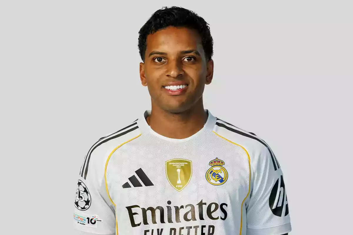 Jugador de fútbol con uniforme blanco del Real Madrid posando y sonriendo frente a un fondo gris claro Jugador de fútbol con uniforme blanco del Real Madrid posando y sonriendo frente a un fondo gris claro